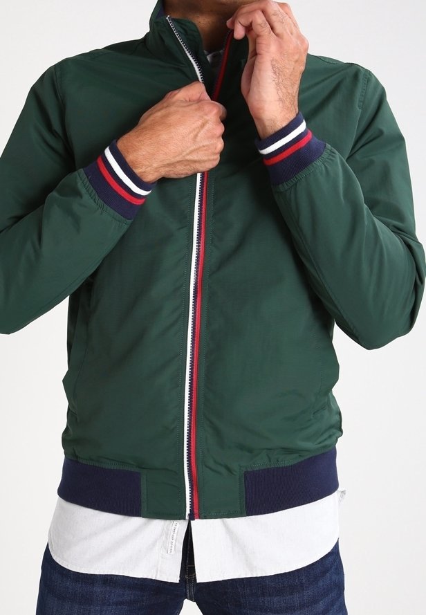 Muž zapínající tmavě zelenou bundu s manžetami a pasem s pruhy v námořnické modři, bílé a červené barvě, přes bílou košili a tmavé džíny.