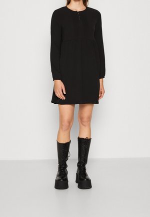 Kobieta ubrana w czarną sukienkę z długim rękawem, do tego czarne platformowe kozaki sznurowane do kolan, stojąca na jednolitym białym tle.