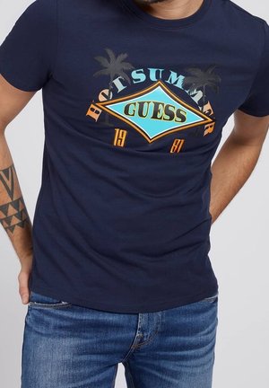T-shirt bleu marine avec le logo "GUESS", des palmiers et le texte "Best Summer 1981" porté par un homme tatoué en jean bleu.