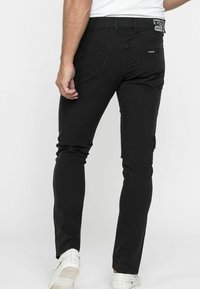 Jeans neri aderenti con una superficie liscia, dotati di tasche posteriori, dettagli cuciti e una cintura con logo, indossati con sneakers bianche.