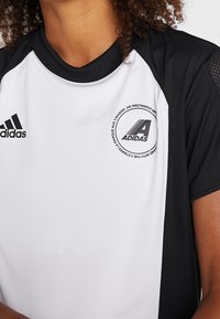 Kraťasový sportovní dres s černobílou barevnou kombinací. Má kulatý výstřih, síťované detaily a kruhové logo Adidas.