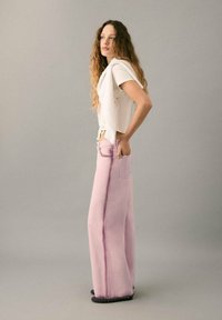 Top alb, scurt, cu mâneci scurte, tăiat deasupra taliei, cu nasturi laterali; pantaloni largi din denim roz cu buzunare frontale și cusătură contrastantă; model în picioare, privit din lateral.