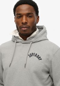Grijze hoodie met een vooraanstaande kangoeroezak, trekkoordcapuchon en zwart "SUPERDRY" logo gestikt op de borst. Zachte stoftextuur.