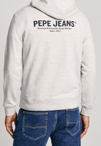 Uomo che indossa una felpa con cappuccio grigio chiaro con scritto sul retro "PEPE JEANS Based on Portobello Road Market Since 1973", abbinata a jeans blu.