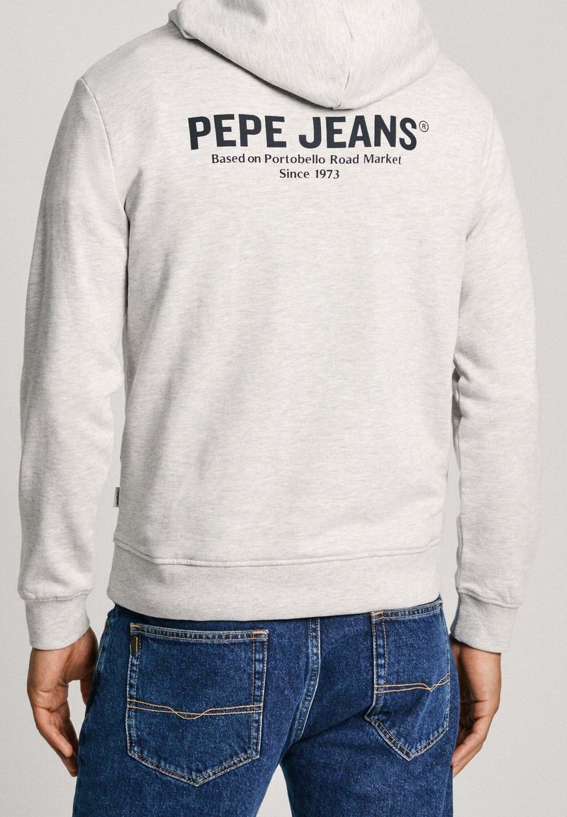 Uomo che indossa una felpa con cappuccio grigio chiaro con scritto sul retro "PEPE JEANS Based on Portobello Road Market Since 1973", abbinata a jeans blu.