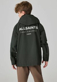 Olivenfarvet vandtæt jakke med hætte. Har et hvidt logo og tekst på ryggen, slankt design og en struktureret tekstur.