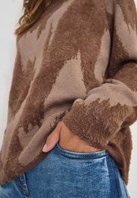 Bruine fuzzy trui met golfpatroon in lichtere bruintinten, voorzien van geribde manchetten en een ronde zoom, gecombineerd met blauwe denim jeans.