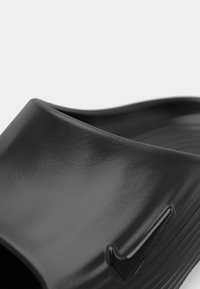 Μαύρο παντόφλι με γυαλιστερή επιφάνεια, με στρογγυλή κορυφή και υφασμάτινη εσωτερική σόλα, διακοσμημένο με μια μικρή μαύρη λεπτομέρεια λογότυπου στο πλάι.