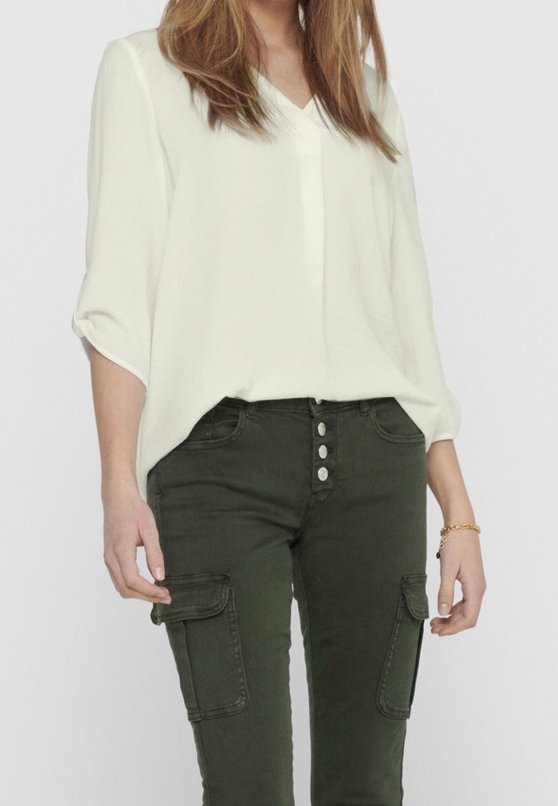 Blusa blanca de cuello en V combinada con pantalones cargo verde oscuro que cuentan con cierre de botones y bolsillos laterales. Tela lisa con un ajuste relajado.