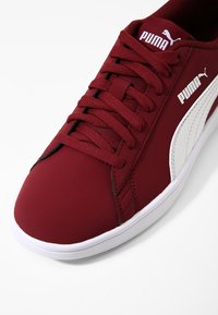 Zapatilla deportiva roja con parte superior de tela suave, detalles en cuero blanco, cordones planos y suela de goma texturizada. Presenta un logo en el costado.