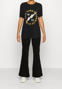 Černé tričko s krátkým rukávem s bílým a zlatým grafickým designem s bleskem, v kombinaci s rozšířenými černými kalhotami a bílými teniskami.