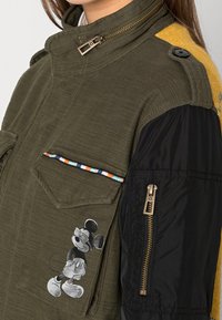 Olive green jacket with zip front, contrasting black sleeves, and two chest pockets, featuring a Mickey Mouse graphic and multicolored accent stitching.