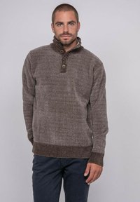 Homme debout les mains dans les poches, portant un pull en maille texturé marron avec col boutonné et un pantalon sombre.
