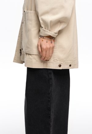 Osoba v béžovej bunde a čiernych nohaviciach s náramkom z korálok s farebnými kameňmi na ľavom zápästí.