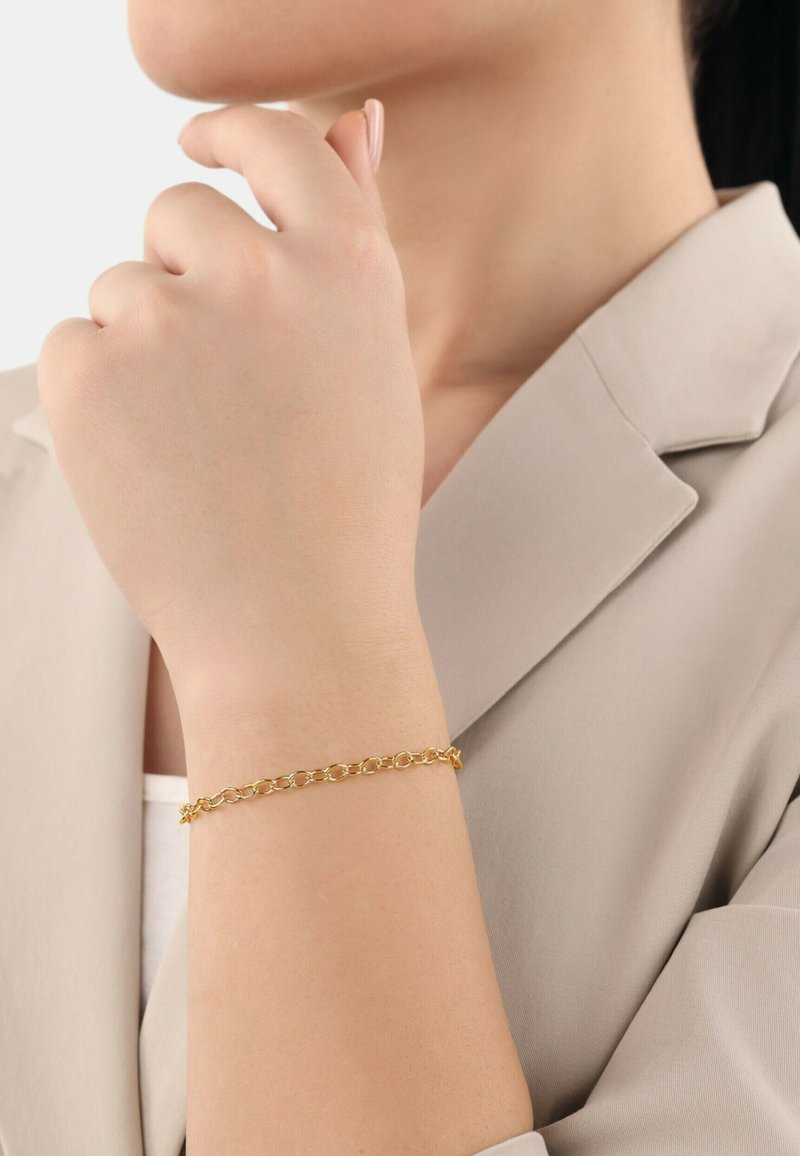 Gold chain bracelet on a wrist, featuring interlocking oval links. Smooth texture with a polished finish, worn over a light beige sleeve.