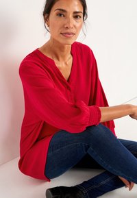 Rode gestructureerde blouse met een knoopsluiting bij de hals, losse mouwen en geplooide manchetten; gecombineerd met donkerblauwe jeans en zwarte schoenen.