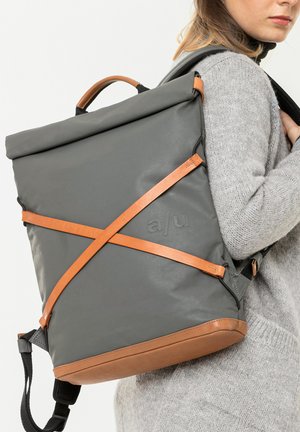 Vrouw met een grijze en bruine roll-top rugzak met gekruiste leren riemen over een lichtgrijze trui.