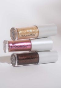 Tre cylindriske kosmetikkflasker med hvite lokk. Den øverste har gullglitter, den midterste har rosa shimmer, og den nederste har mørkebrun shimmer.