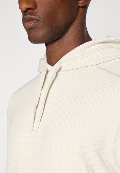Ljust beige huvtröja med texturerad ribbad tyg, justerbara dragsnören och minimala sömnadsdetaljer på axeln.