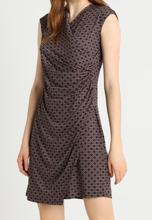 Robe sans manches brun foncé avec un motif circulaire en chaîne de couleur beige clair, cintrée à la taille, portée par une personne aux longs cheveux roux.