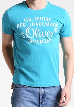 Mężczyzna ubrany w jasnoniebieski krótki rękaw t-shirt z białym napisem "s.Oliver Customized" oraz wyblakłe niebieskie dżinsy na jednolitym tle.