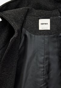 Interno di un cappotto di lana nero con una fodera nera liscia. L'etichetta attaccata riporta la scritta "Koton", taglia EUR 36, con dettagli di cucitura minimi.