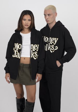 Twee personen dragen zwarte hoodies met de tekst "Money Talks?"; de vrouw draagt een wit crop top, een groene rok en zwarte laarzen; de man draagt een zwarte broek.
