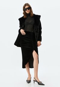 Zwarte nep bontjas over een doorschijnende zwarte top en een gerimpelde fluwelen rok met een zijsplit. Puntige zwarte hakken maken de look compleet.