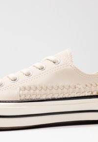 Baskets en toile beige avec détails tissés sur les côtés, œillets argentés et semelle extérieure en caoutchouc blanc surélevée avec une bande noire.