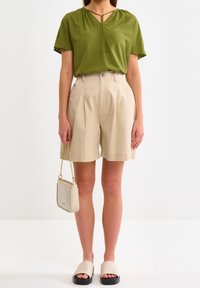 Haut vert olive à col en V avec détail noué, short plissé taille haute beige, sandales slides crème et petit sac bandoulière crème.