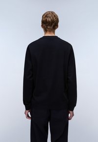 Osoba o krótkich falowanych włosach, ubrana w gładką czarną koszulę z długim rękawem i czarne spodnie, stojąca tyłem na neutralnym tle.