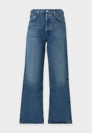 Une femme porte une chemise blanche, un haut gris, un jean bleu, des ballerines bordeaux et un sac à main assorti, sur un fond blanc uni.