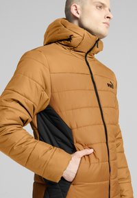 Giacca a vento marrone con chiusura a zip, cappuccio e pannelli laterali neri. Presenta una texture trapuntata e un logo sul petto.
