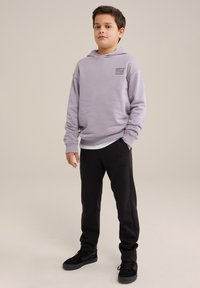 Svetložltý mikina s kapucňou a predným vreckom, s malou grafikou na hrudi, spárovaná s čiernymi teplákmi a čiernymi teniskami.