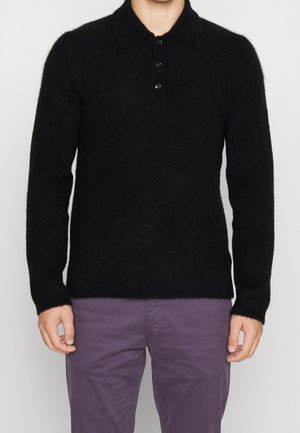 Homme portant un polo à manches longues noir en tissu duveteux et un pantalon violet, debout devant un fond clair uni.