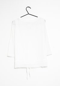 Blusa bianca semi-trasparente con maniche a tre quarti, scollatura rotonda e dettaglio con fiocco sull'orlo inferiore, esposta su una gruccia nera.