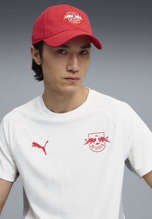 Młody mężczyzna w czerwonej czapce RB Leipzig i białej koszulce Puma z logo RB Leipzig na jednolitym szarym tle.