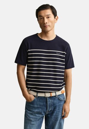 Mann trägt ein marineblaues kurzärmeliges Hemd mit weißen horizontalen Streifen, blaue Jeans und einen hellbeigen gewebten Gürtel, steht mit einer Hand in der Hosentasche.