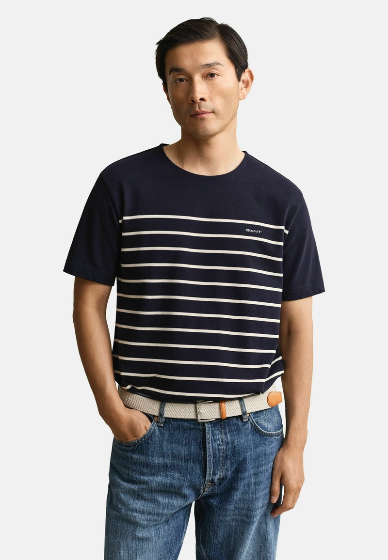 Homme portant une chemise à manches courtes bleu marine avec des rayures horizontales blanches, un jean bleu, et une ceinture tissée beige clair, debout avec une main dans la poche.