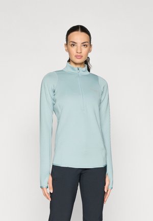 Haut zippé à manches longues bleu clair avec un motif texturé et des trous pour les pouces aux poignets. Associé à un pantalon noir, il présente une coupe ajustée.