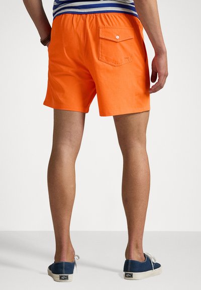 Oransje shorts med baklomme; elastisk midje, tettsittende design, kombinert med marineblå sko. Glatt stoffstruktur og uformell stil.