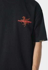 Zwarte t-shirt met korte mouwen, voorzien van een rode gestreepte Pegasus-logo en de tekst "PAS DE MONACO" op de linkerborst, gedragen door een persoon.