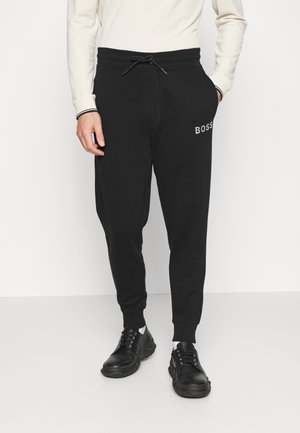 Man draagt zwarte joggers met de tekst "BOSS" op de dij, een wit shirt met lange mouwen en zwarte schoenen, staand tegen een effen witte achtergrond.