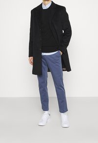 Mann in einem schwarzen Überzug, schwarzem Pullover, hellblauem Hemd, blauen Hosen und weißen Sneakers, der mit einer Hand in der Tasche steht.