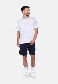 Witte katoenen t-shirt, ronde hals, met een klein logo in goud. Gecombineerd met marineblauwe shorts, witte sokken en witte sneakers.