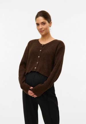 Cardigan marron à côtes avec un décolleté en V, comportant cinq grands boutons sur le devant et des manches légèrement raccourcies. Associé à un pantalon noir.
