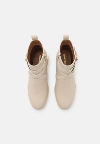 Bottes beige à la cheville en daim lisse. Dotées de brides croisées et d'une fermeture éclair latérale. Accents en métal doré et semelle plate.