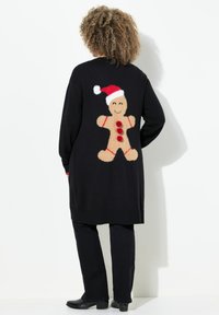 Persoon met krullend haar in een lange zwarte cardigan met een grote glimlachende peperkoeken man met een rode kerstmanhoed op de achterkant.
