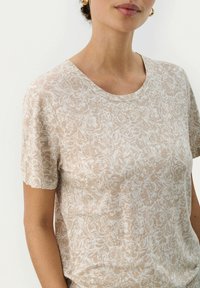 T-shirt beige à manches courtes avec un motif floral, encolure ronde et texture douce. Présente une coupe décontractée et un design casual.