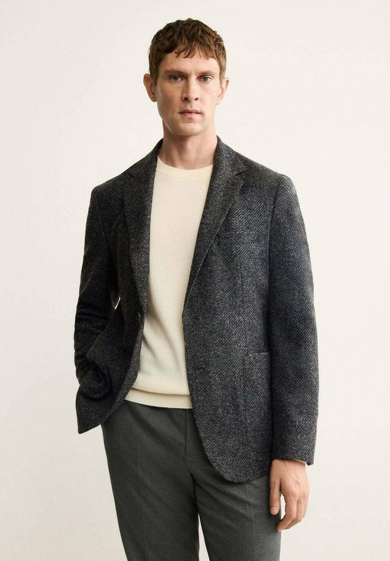 Blazer grigio scuro a trama texturizzata con un sottile motivo a spina di pesce, abbinato a un maglione beige chiaro, completo di classica rever a punta.